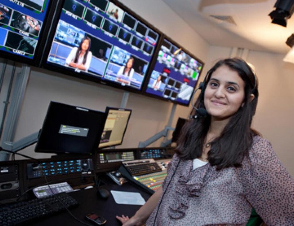 Image of a student wearing a set of headphones with a microphone. They are standing in front of a row of TV monitors and other audiovisual equipment.