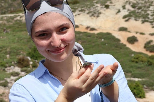 A smiling woman wearing a light gray headscarf and sunglasses holds a small turtle in her hands outdoors, with hills and greenery in the background.