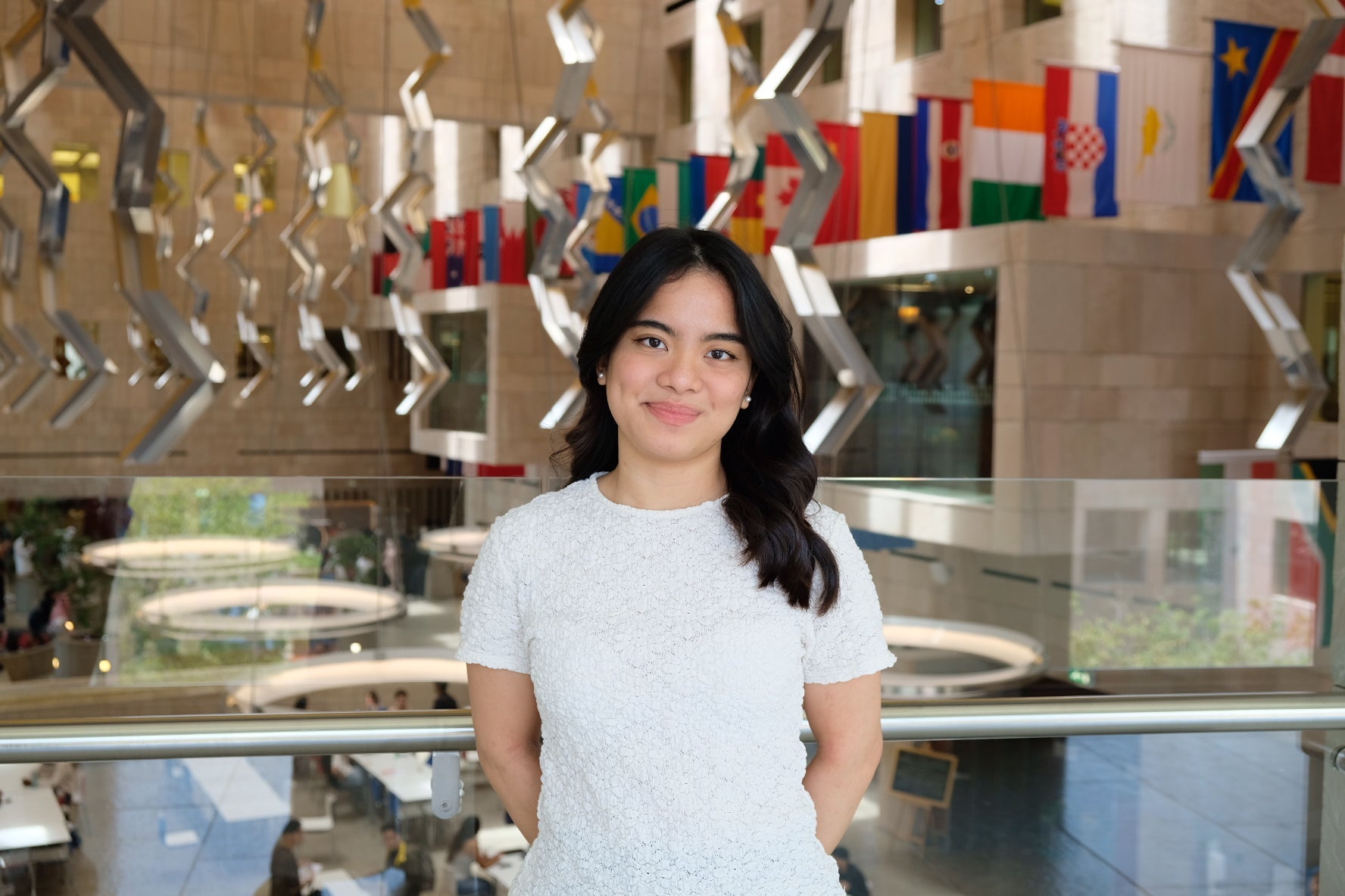 A young woman with long dark hair and a white textured top stands smiling indoors, with international flags and modern architectural decor in the background.