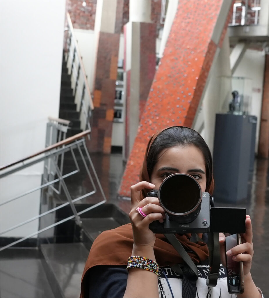 A person wearing a brown headscarf and colorful bracelets holds a camera up to their face, taking a photo indoors near a staircase and modern architectural elements.