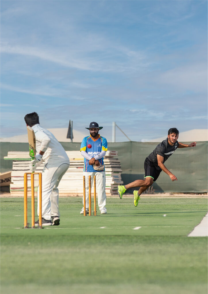 A cricket bowler in black delivers the ball as a batsman in white prepares to hit, while an umpire watches closely from behind the stumps on a green pitch under a blue sky.