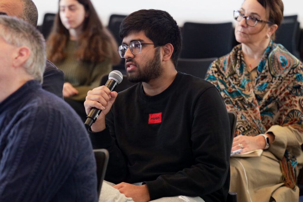A young man with glasses holds a microphone and speaks during an event, wearing a red name tag that says Hugo. People are seated around him, listening attentively.