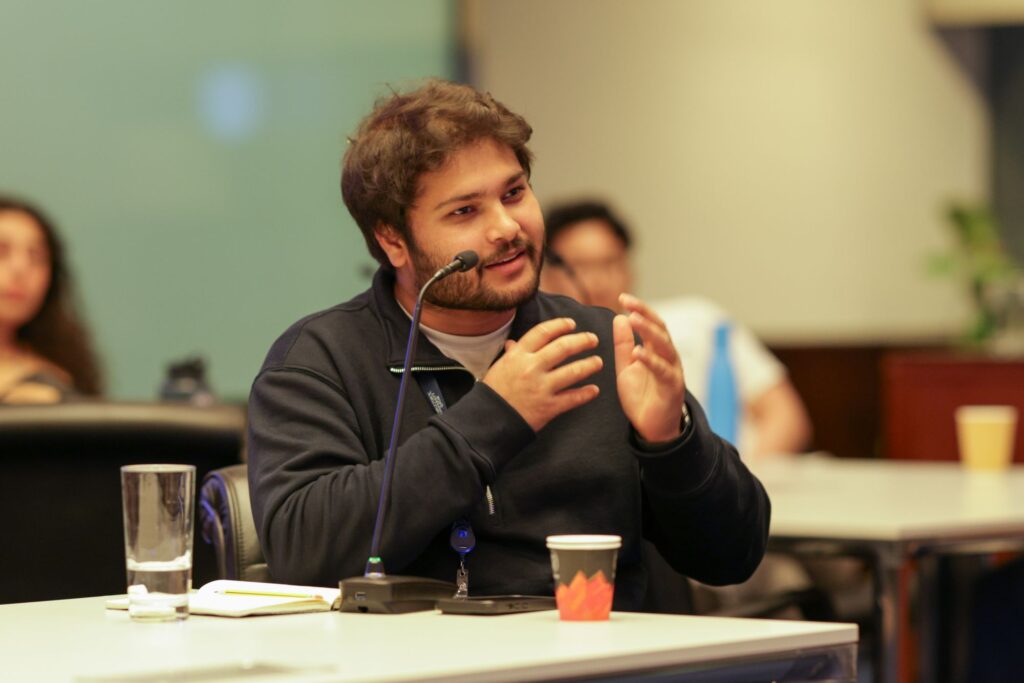 A man with dark hair and a beard sits at a table, speaking into a microphone and gesturing with his hands. A notepad, glass of water, and paper cup are in front of him. Other people are blurred in the background.