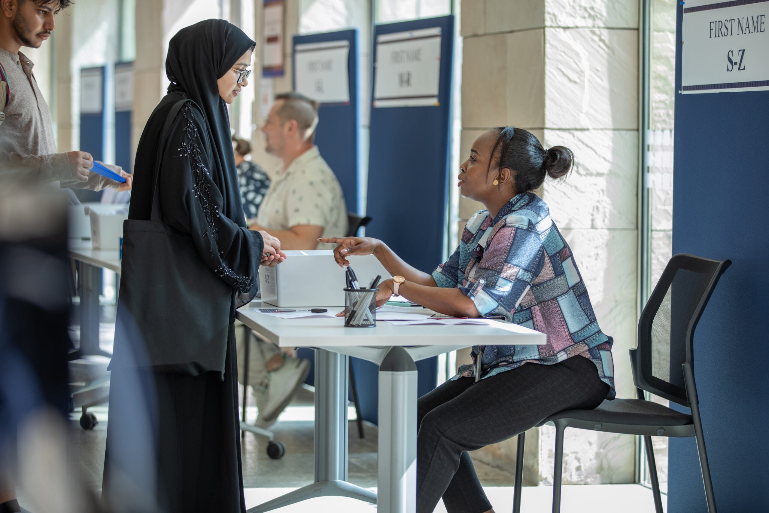 A woman in a hijab stands at a registration desk, speaking with a seated woman who is gesturing. Other people sit at desks in the background, and signs with last name ranges are visible on the partitions.