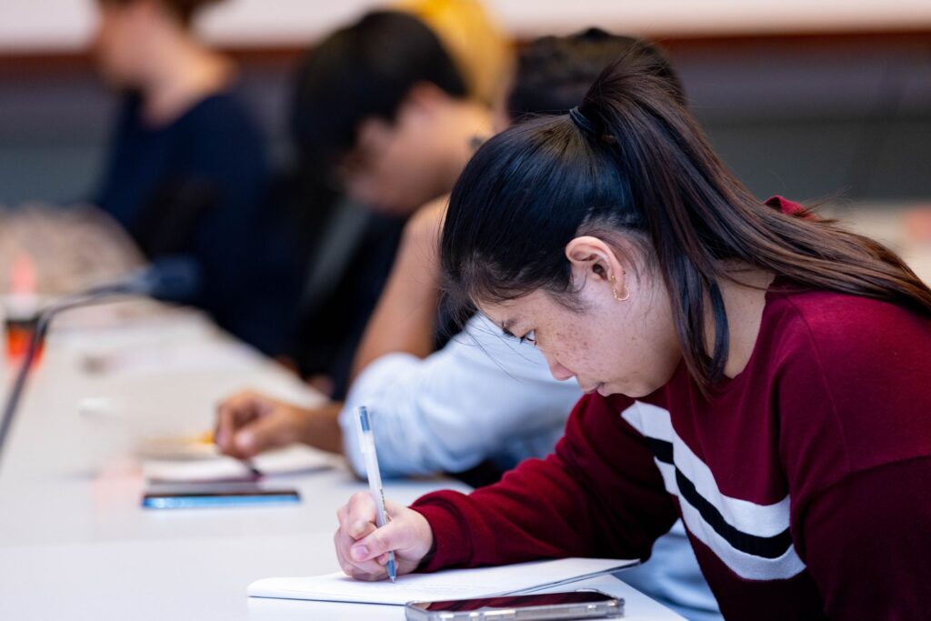 A young woman in a maroon sweater writes in a notebook at a desk, surrounded by other students who are also focused on their work in a classroom setting.