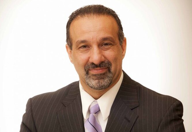 A man with short dark hair, a trimmed beard, and mustache, wearing a dark pinstripe suit, white shirt, and light purple tie, smiles slightly in front of a plain light background.
