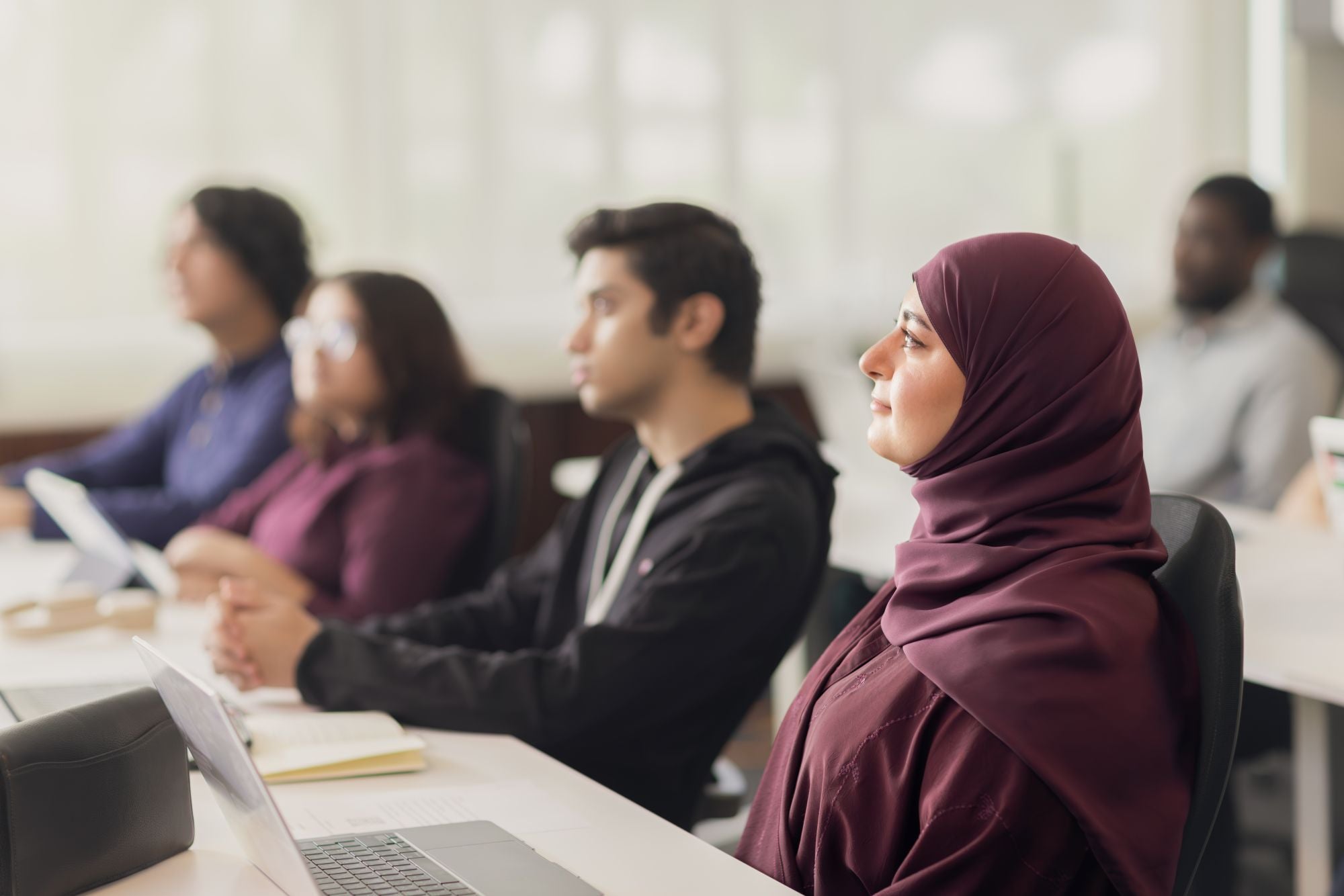 A group of diverse adults sit at desks in a bright classroom, attentively listening. A woman in a maroon hijab sits in the foreground, focused, with others blurred in the background. Laptops and notebooks are on the desks.