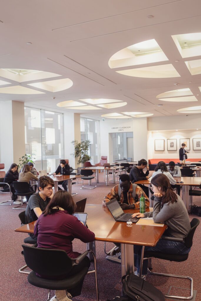 Students sit at tables in a spacious, brightly lit library room, working on laptops and studying. Several groups and individuals are focused on their tasks, with large windows and modern decor in the background.