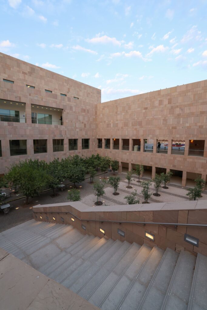 A modern courtyard with neatly arranged young trees, surrounded by a tan stone building with large windows and a wide staircase leading down to the courtyard, under a blue sky with light clouds.