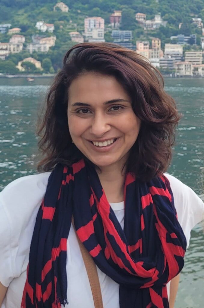 A woman with wavy brown hair smiles in front of a lake. She is wearing a black and red scarf over a white top. In the background, there are hills and buildings along the shoreline.