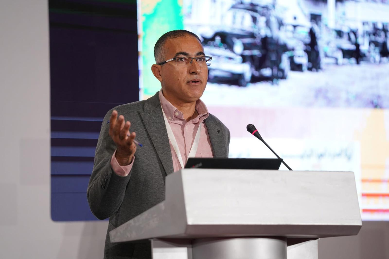 A man wearing glasses and a gray blazer speaks at a podium with a microphone. A colorful, partially blurred screen with images and text is visible in the background.