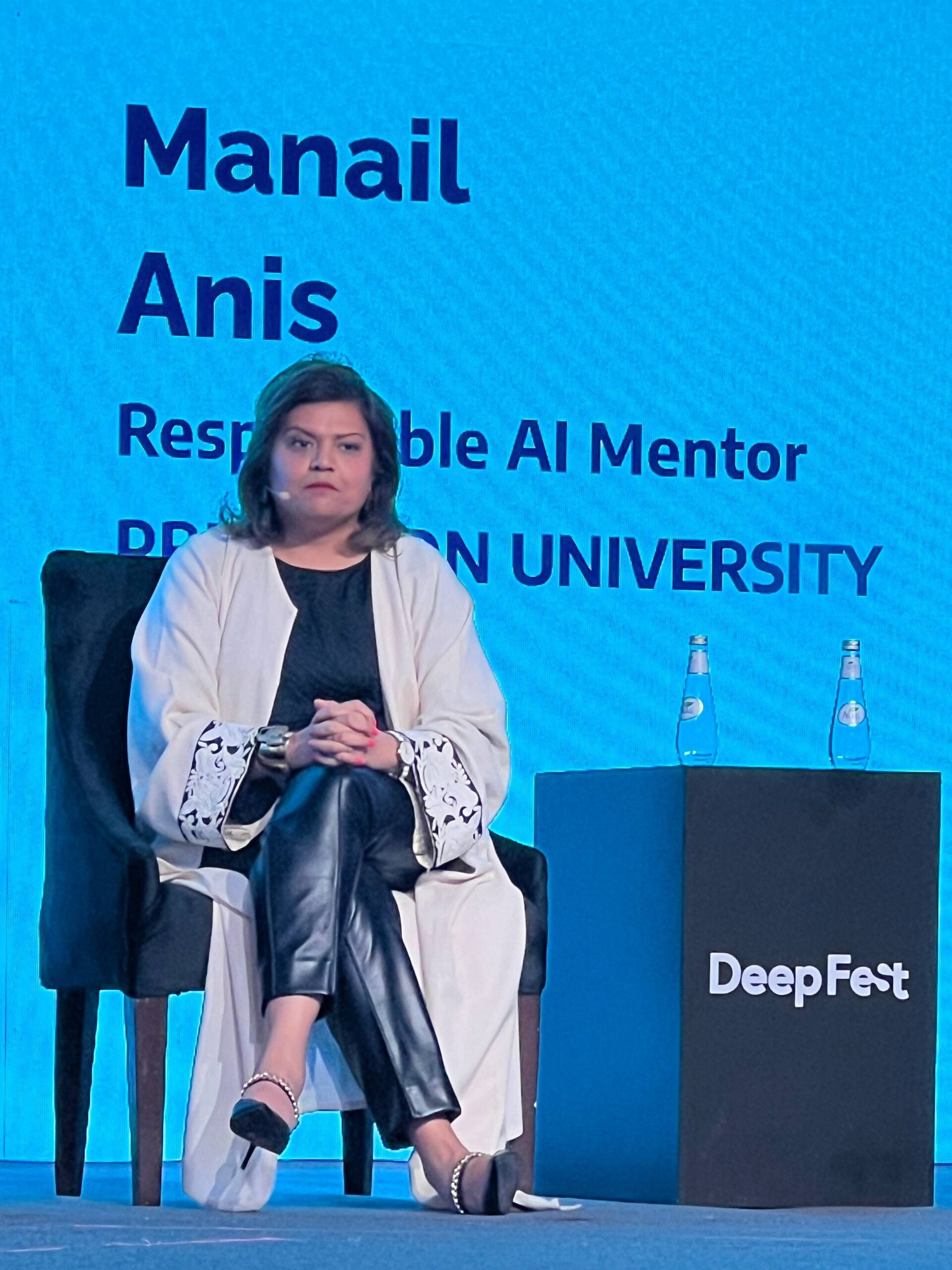 A woman sits on a stage chair, hands clasped, in front of a blue screen displaying the name Manail Anis, Responsible AI Mentor, Princeton University at DeepFest. Two water bottles are on a table beside her.
