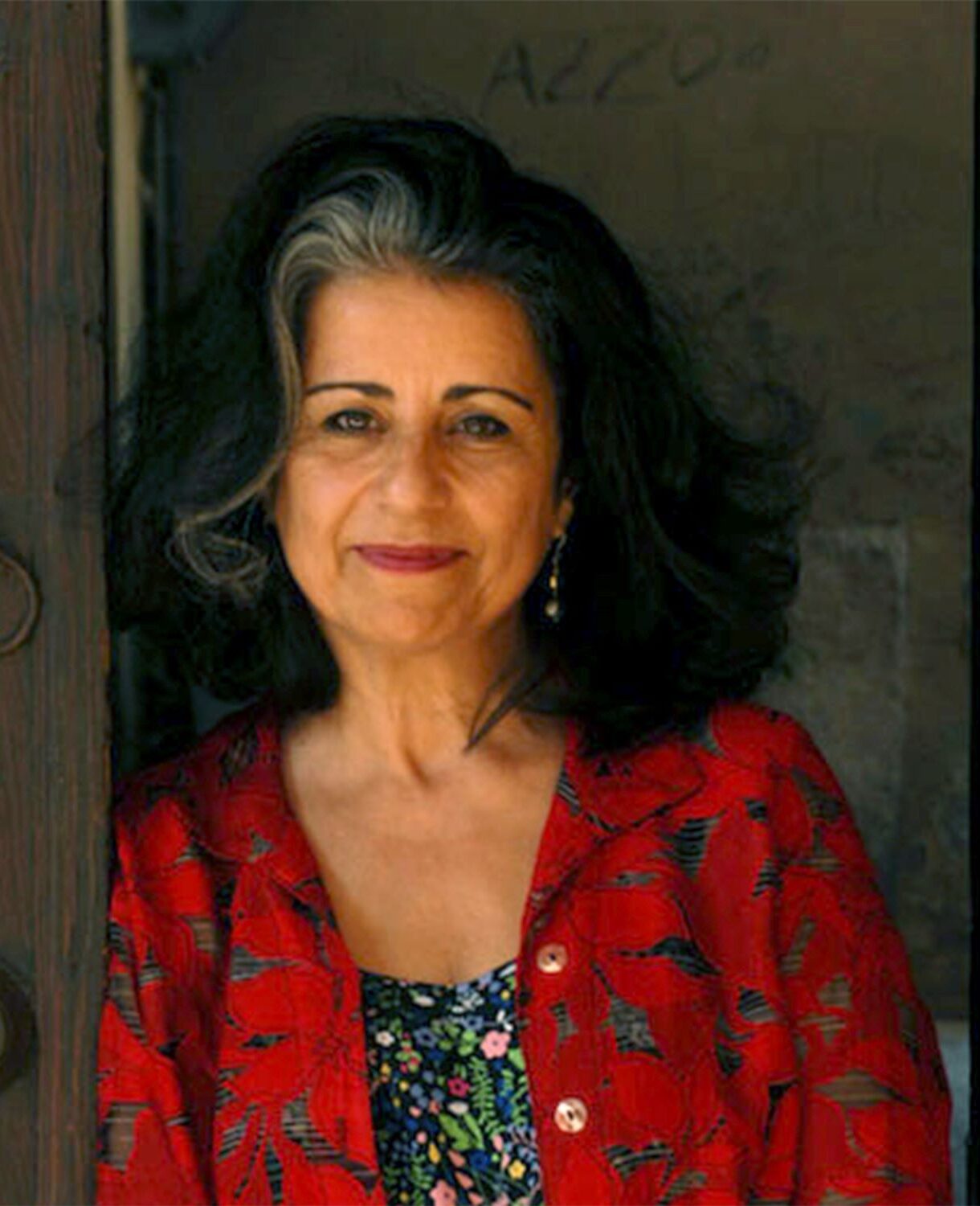 A woman with dark, shoulder-length hair featuring a white streak stands by a wooden door. She wears a red patterned jacket over a floral top and smiles gently at the camera. The background is dim and slightly blurred.