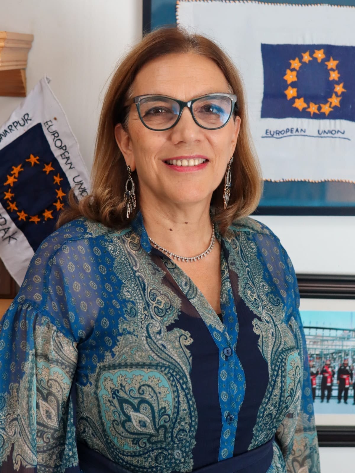 A smiling woman with glasses and shoulder-length brown hair wears a blue patterned blouse and silver jewelry, standing in front of European Union flags and framed pictures.