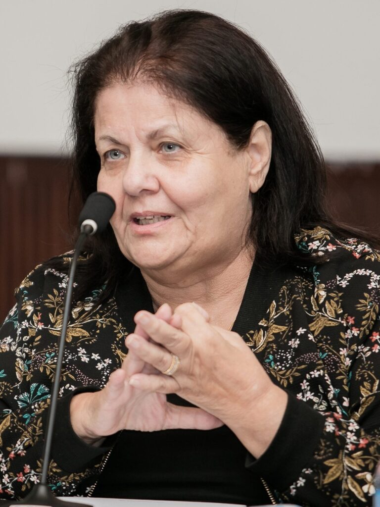 A woman with dark hair wearing a black floral-patterned jacket speaks into a microphone. She is gesturing with her hands and appears to be in a discussion or giving a presentation.