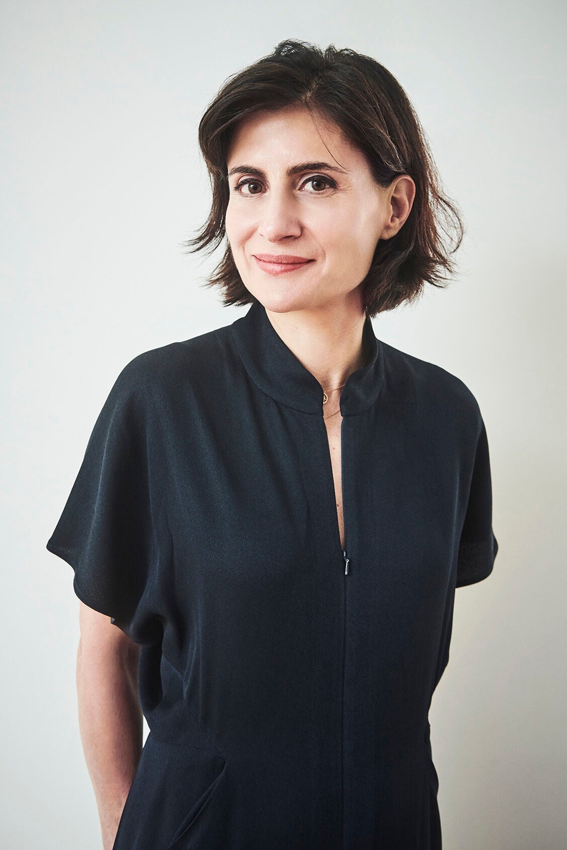 A woman with short dark hair wearing a dark, short-sleeved dress stands against a light background, smiling softly at the camera.