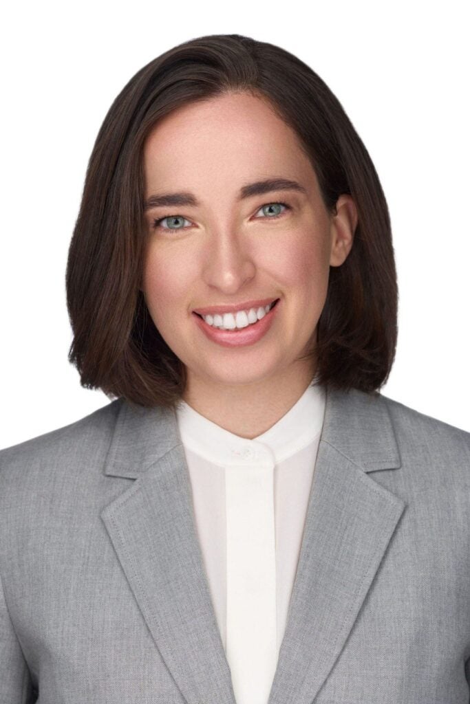 A person with straight brown hair and blue eyes is smiling, wearing a light gray blazer over a white button-up shirt, posed against a plain white background.