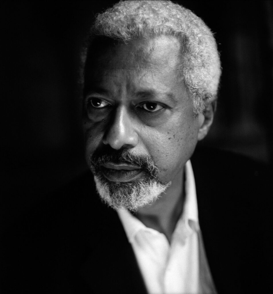 A grayscale portrait of an older man with short curly gray hair, a mustache, and a goatee. He is wearing a white shirt and a dark jacket, looking thoughtfully to the side against a dark background.