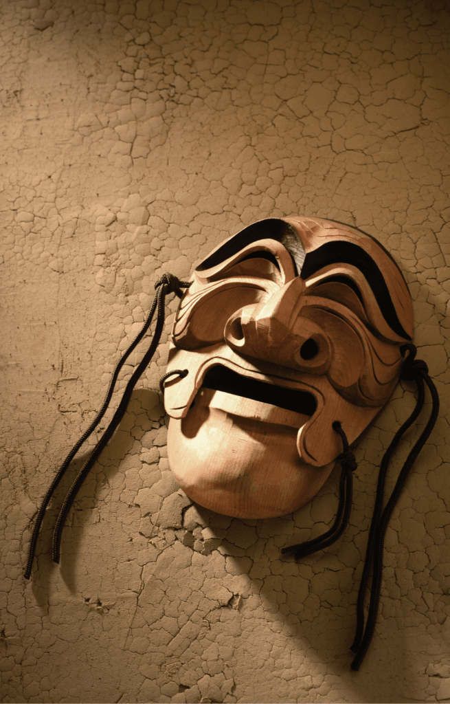 A wooden theatrical mask with black accents and cord ties rests on a cracked, textured surface, lit by warm, dramatic light from the side.