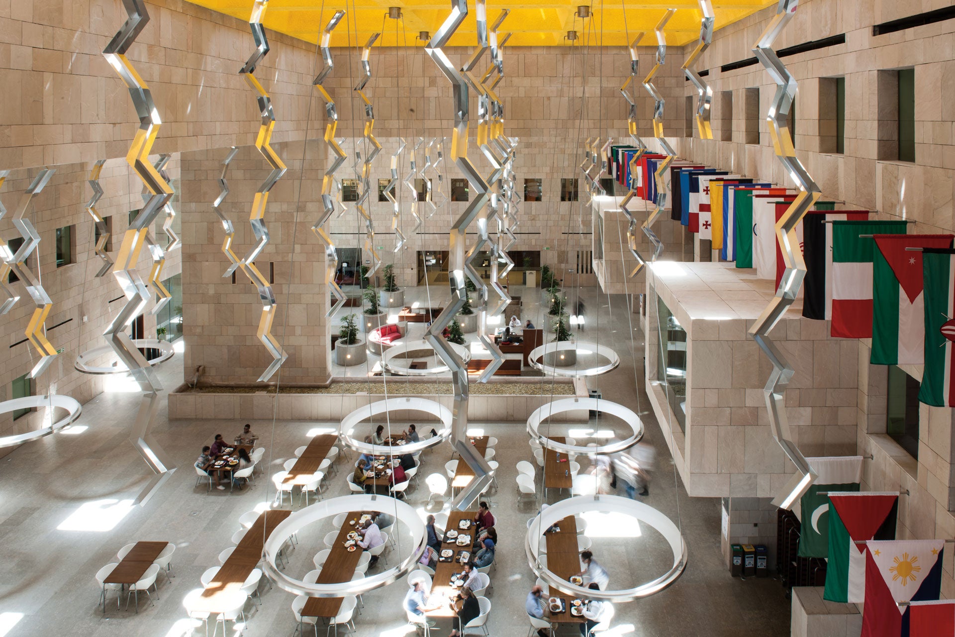 A spacious, modern atrium with tall beige stone walls, hanging metallic zig-zag sculptures, round light fixtures, and multiple international flags. People sit at long wooden tables below, some eating or talking.