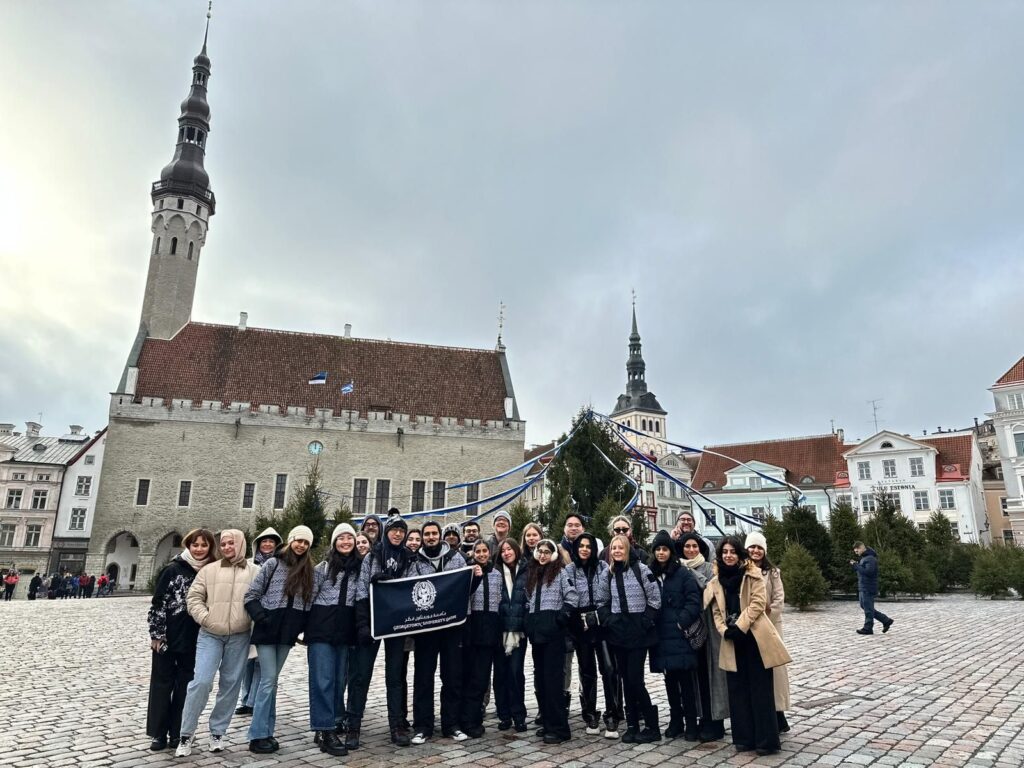 A group of people poses together in a cobblestone square with a historic building and tall spire in the background. Some hold a black flag with a white emblem. The sky is overcast, and evergreen trees line the area.