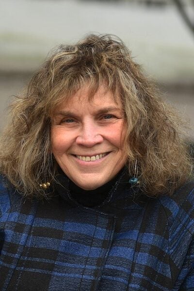 A woman with curly, shoulder-length hair smiles at the camera. She is wearing a dark plaid jacket and dangling earrings, and she is outdoors.