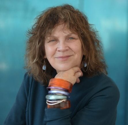 A woman with curly brown hair smiles gently, resting her chin on her hand. She wears a dark top, colorful chunky bracelets, and drop earrings, with a teal-blue background behind her.