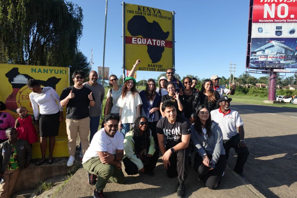 A group of people pose and smile in front of a yellow sign that marks the equator in Nanyuki, Kenya. The sign displays a map of Africa and the word Equator. Some local children are also present on the left.