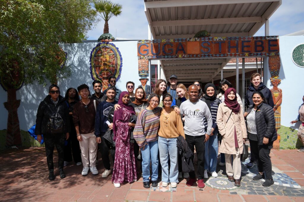 A diverse group of people pose and smile in front of the colorful entrance to Guga S’thebe cultural center, with vibrant murals and outdoor decorations visible behind them on a sunny day.