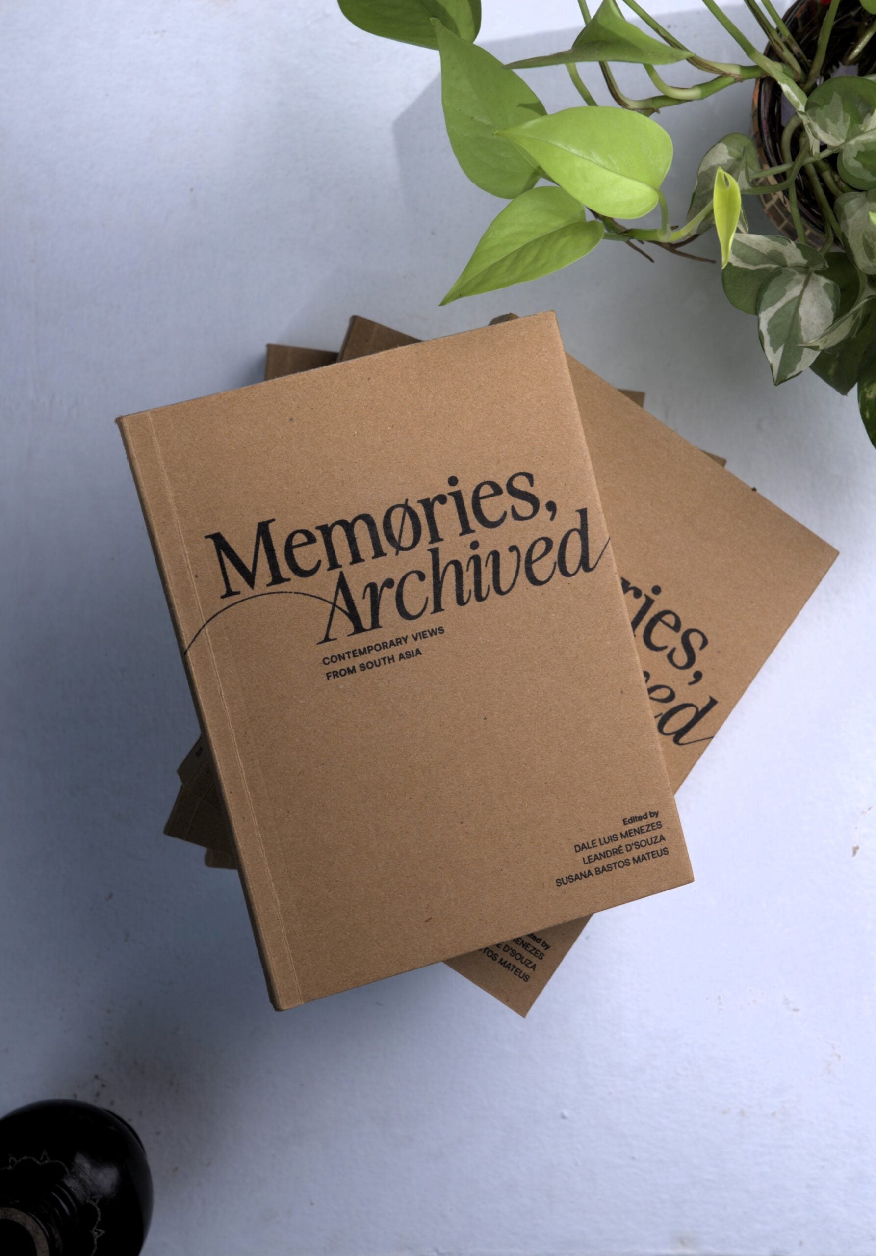 A stack of brown books titled Memories, Archived: Contemporary Views from South Asia sits on a light surface next to a green potted plant. The cover features simple, modern typography.