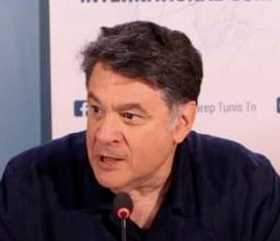 A middle-aged man with dark hair speaks into a microphone at a press conference, seated in front of a light blue and white background with partially visible text.