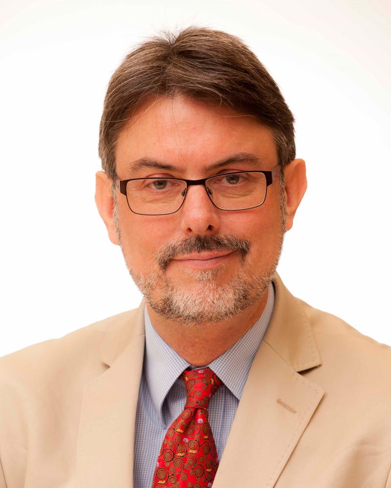 A man with short brown hair, glasses, and a beard, wearing a beige suit jacket, a blue shirt, and a red patterned tie, is smiling slightly against a plain white background.