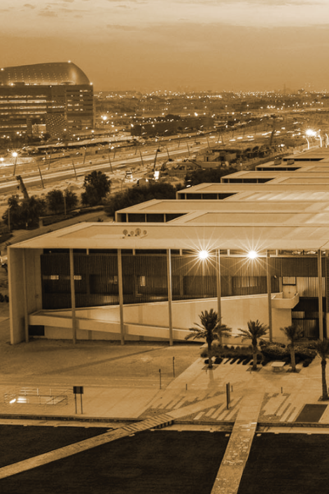 Sepia-toned image of a modern building with large windows and palm trees in front, set against a cityscape with a prominent curved glass structure and illuminated roads at dusk.