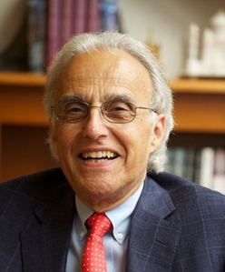 An older man with gray hair and glasses smiles while wearing a blue suit, light blue shirt, and red tie. Bookshelves are visible in the blurred background.