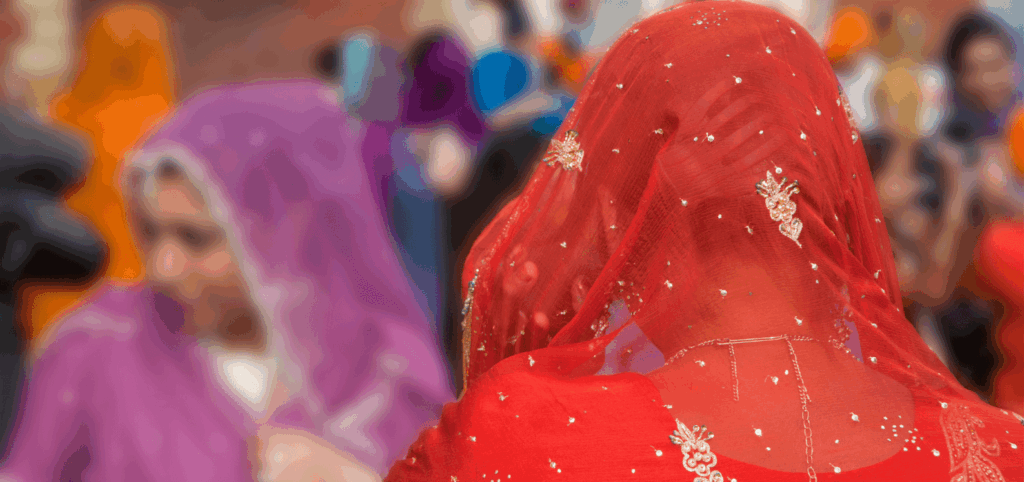 Two women wearing embroidered veils, one in red facing away from the camera and one in purple, stand among a blurred crowd in colorful attire, possibly at a cultural or religious event.