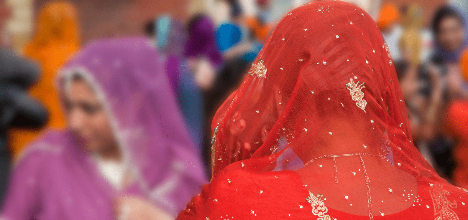 Two women wearing embroidered veils, one in red facing away from the camera and one in purple, stand among a blurred crowd in colorful attire, possibly at a cultural or religious event.