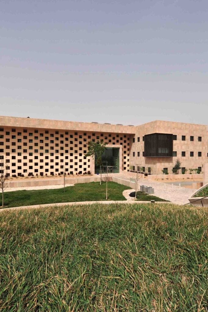 A modern building with a grid-like pattern of square windows and tan stone facade, surrounded by green grass and a paved walkway under a clear sky.