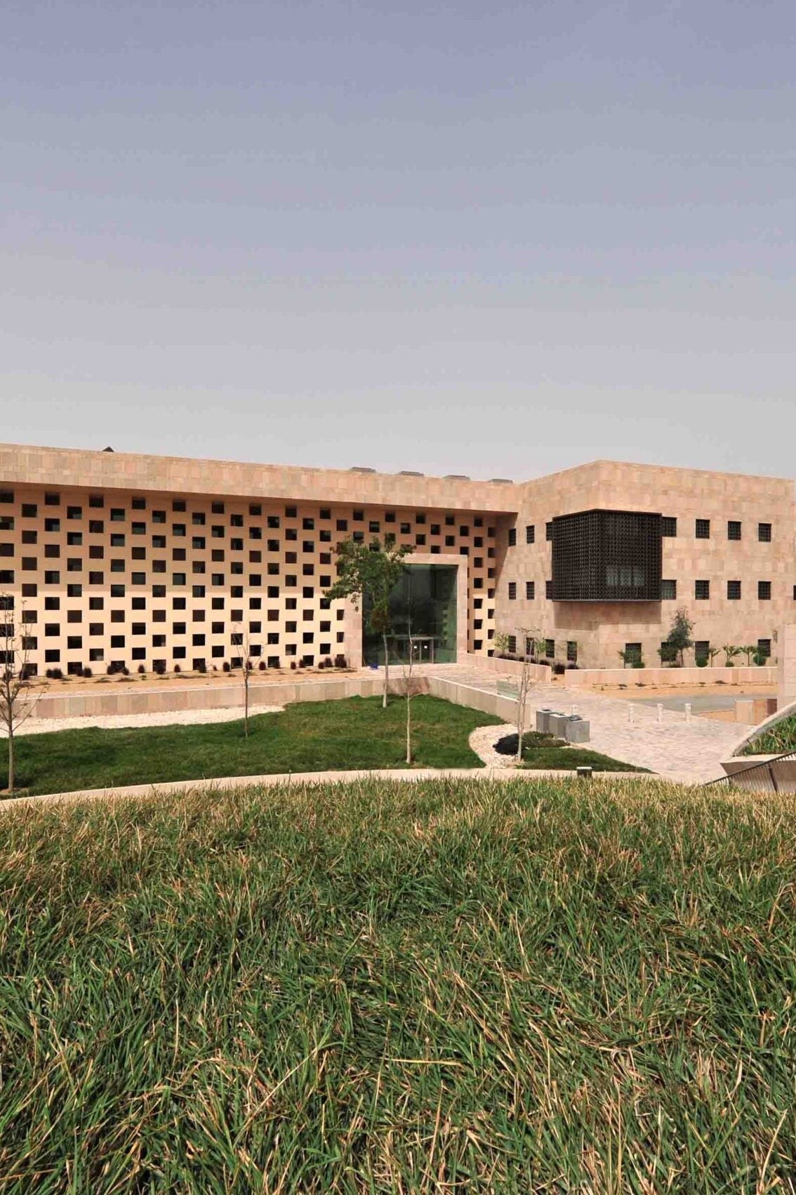 A modern building with a grid-like pattern of square windows and tan stone facade, surrounded by green grass and a paved walkway under a clear sky.