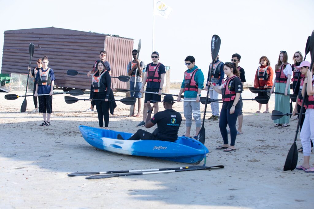 A group of students wearing life jackets hold kayak paddles on the beach as an instructor, seated in a blue kayak, gives a safety briefing.