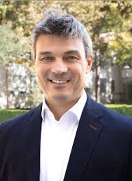 A man with short gray hair, wearing a white shirt and a dark blazer, smiles while standing outdoors in a sunlit park with green trees in the background.