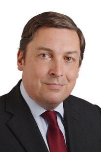 A man with short brown hair wearing a dark suit, light blue shirt, and red tie is facing the camera and smiling slightly against a white background.