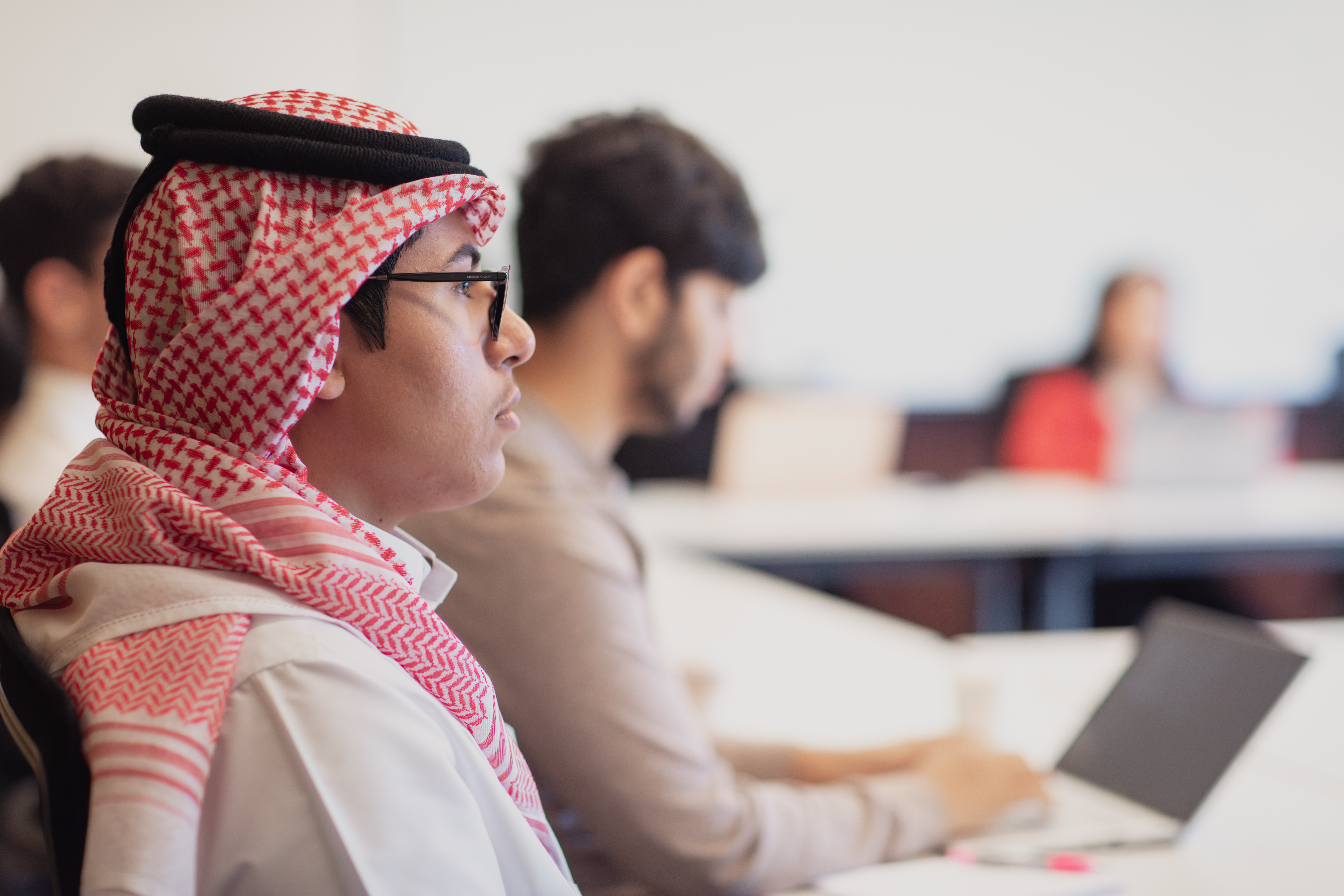 Side profile of a man wearing a traditional headscarf and glasses, seated in a meeting room and listening attentively, with other participants working on laptops in the background.