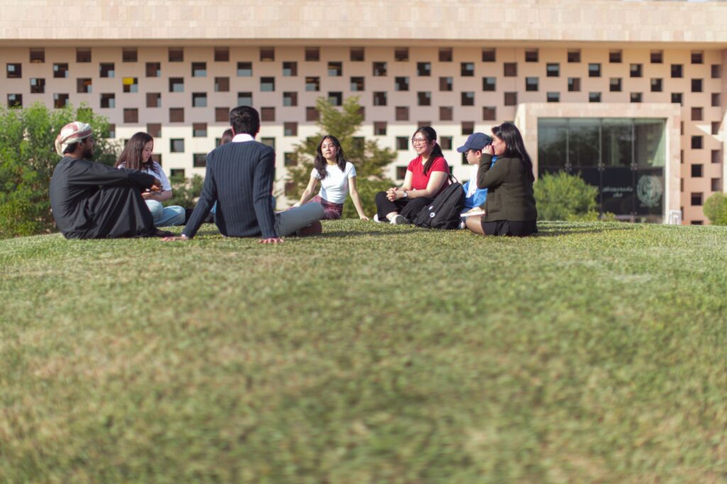 A group of seven people sit in a circle on a grassy lawn, talking and smiling, with a modern building featuring a patterned wall in the background.