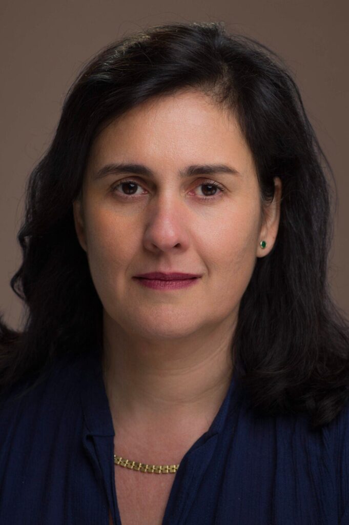 A woman with medium-length dark hair and light skin looks directly at the camera. She is wearing a dark blue top, a gold chain necklace, and small green stud earrings, with a neutral brown background.