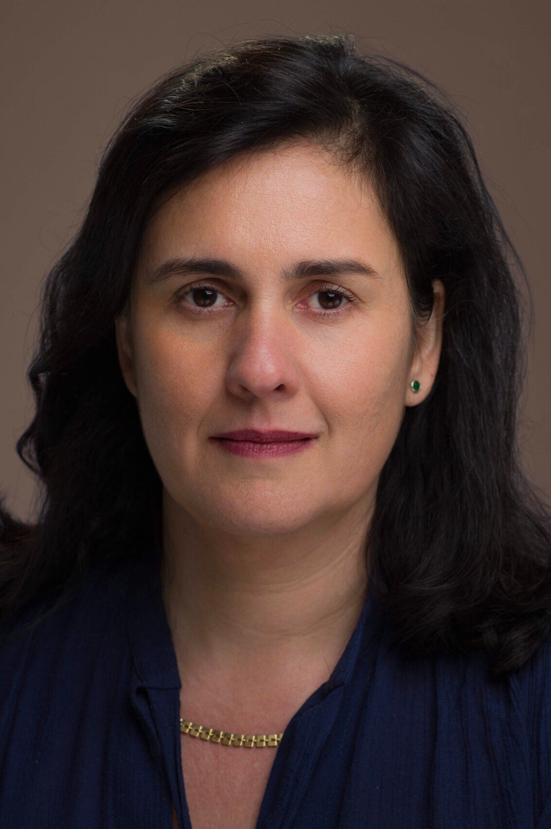 A woman with medium-length dark hair and light skin looks directly at the camera. She is wearing a dark blue top, a gold chain necklace, and small green stud earrings, with a neutral brown background.