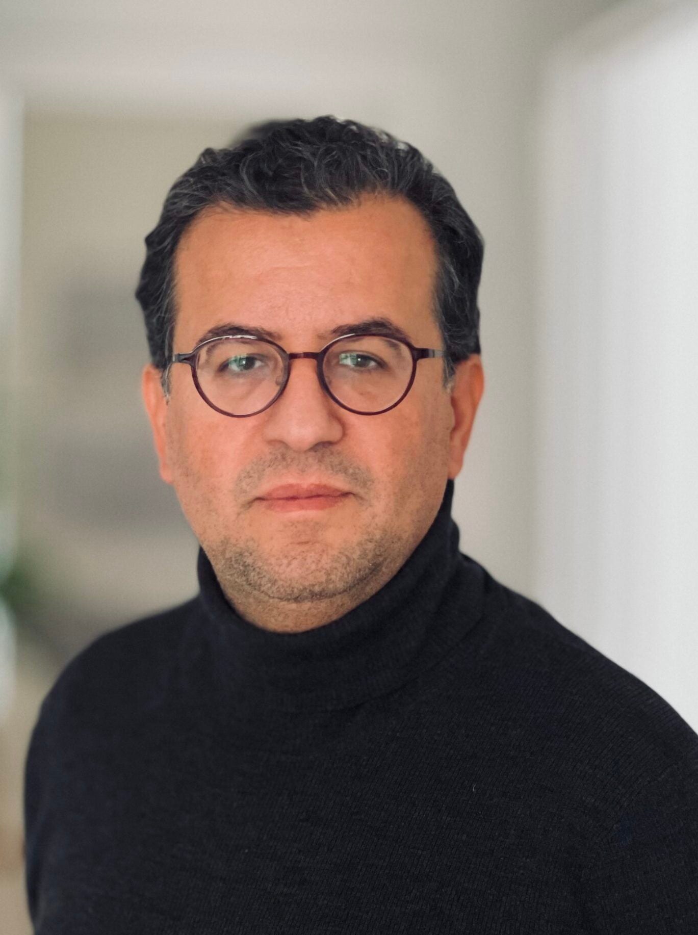 A man with short dark hair, glasses, and light stubble wears a black turtleneck sweater. He is looking at the camera with a neutral expression against a softly blurred indoor background.
