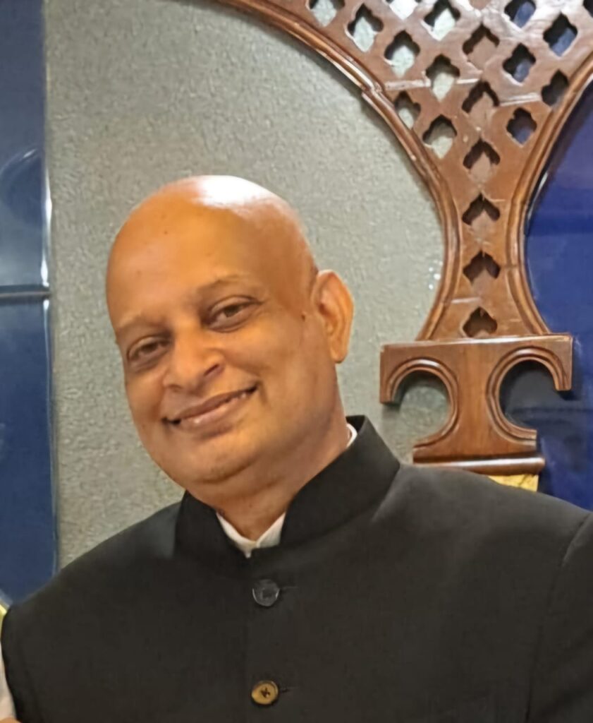 A smiling man with a bald head, wearing a black suit jacket, stands in front of a decorative wooden panel with geometric patterns.