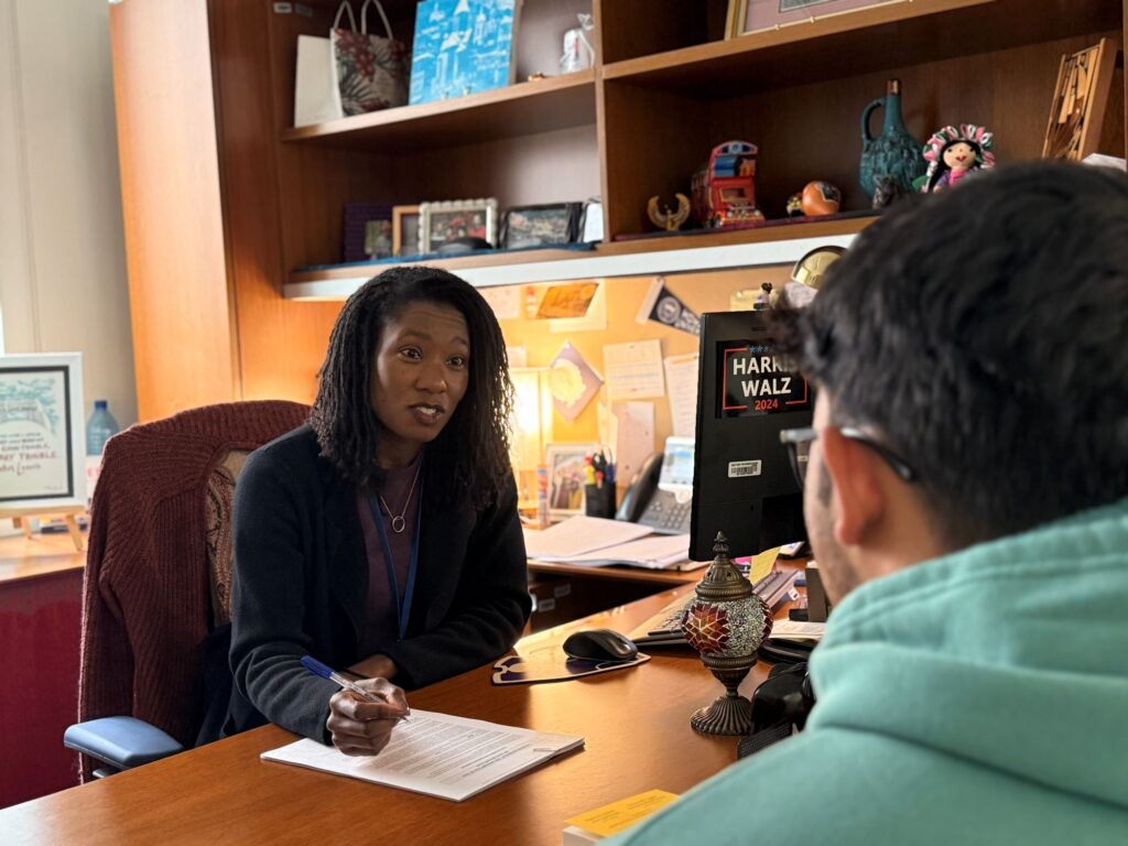 A woman sits at a desk in an office, holding a pen and talking to a person in a green hoodie who is seated across from her. Papers, a computer monitor, and various decorations are visible on the desk and shelves.