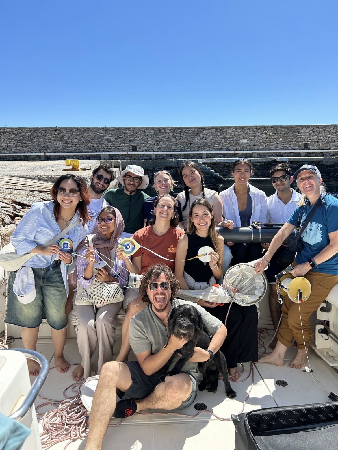 A group of thirteen people, smiling and posing together on a boat under a clear blue sky. They are holding decorative items and one person in front is holding a black dog. There is a stone wall in the background.