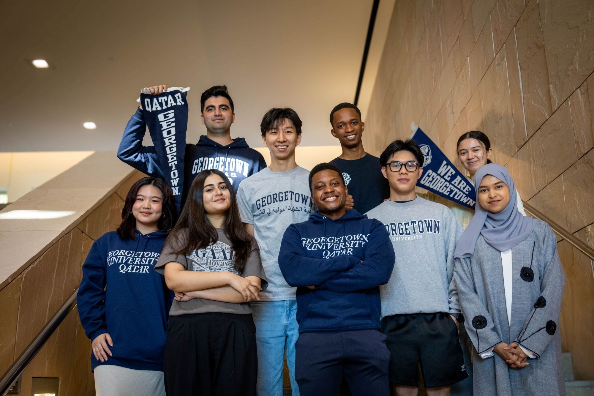 Group of eight Georgetown University in Qatar students pose on an indoor staircase, wearing Georgetown/GU-Q sweatshirts and T-shirts while two hold “Qatar Georgetown” and “We Are Georgetown” banners.